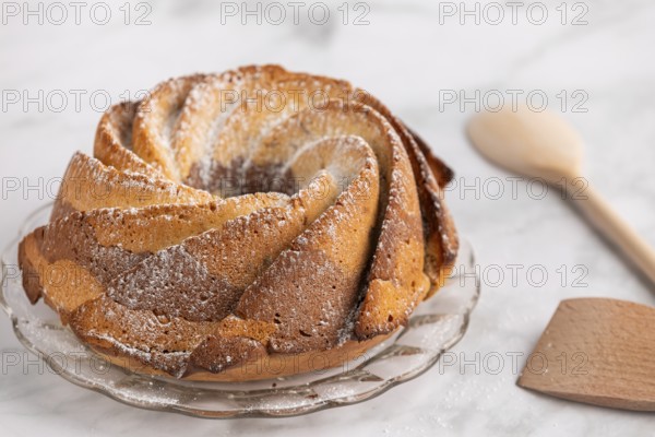 Fresh marble bundt cake, cake with icing sugar next to a rustic wooden spoon on a light-coloured background