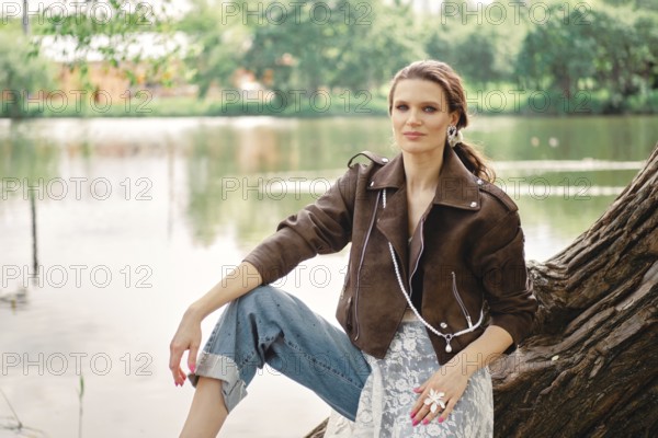 A woman wearing a brown jacket and light blue jeans sits on a tree trunk by a peaceful lake. The sunlight enhances her elegant look amidst the greenery surrounding the water