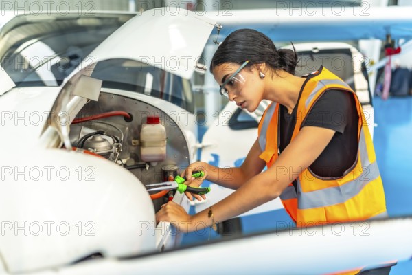 Young woman aircraft mechanic wearing safety glasses and high visibility vest using pliers while working on a small airplane engine inside hangar