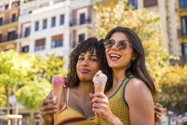 Two smiling girls are eating ice cream cones, hugging each other in a city center during a sunny summer day