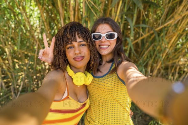 Two happy young women with headphones and sunglasses taking a selfie in a bamboo forest, one of them showing the peace sign
