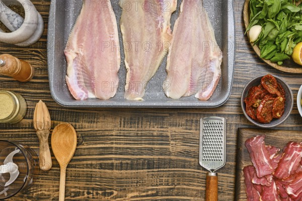 Fresh fish fillets are laid out on a baking tray, surrounded by various ingredients including herbs, lemon, and spices. Wooden utensils and bowls add a rustic touch to the kitchen workspace