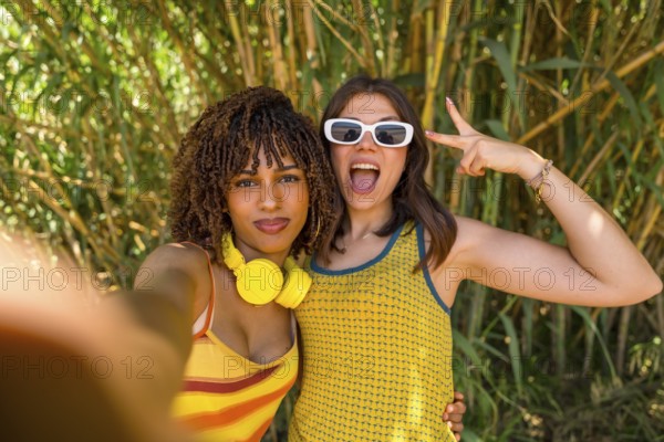 Two girls with headphones and sunglasses taking a selfie in a bamboo forest, making victory signs and funny faces