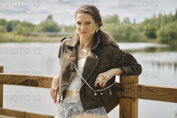 A model stands confidently near a tranquil water body, showcasing a fashionable outfit complete with a brown suede jacket