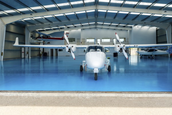 White twin engine airplane parked inside a spacious hangar, featuring a bright blue floor and surrounded by smaller aircraft