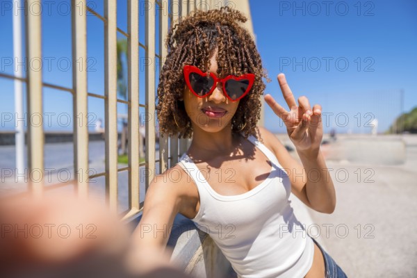 Young woman with heart shaped sunglasses takes a selfie while making the peace sign, leaning against a fence with a beach in the background