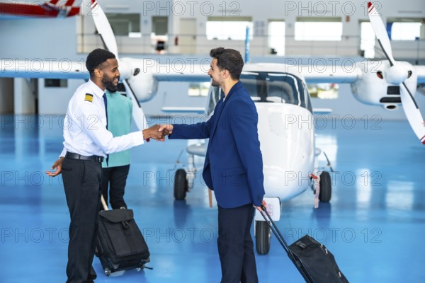 African pilot shaking hands with a businessman inside an aircraft hangar, both holding trolley bags, ready for a private flight
