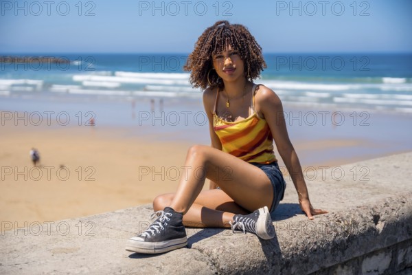 Stylish young woman wearing casual summer clothes sits on a beach wall, enjoying the ocean view during her vacation