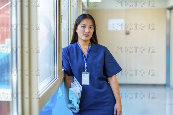 Confident young medic holding a clipboard and wearing blue scrubs is standing next to a window in a hospital corridor