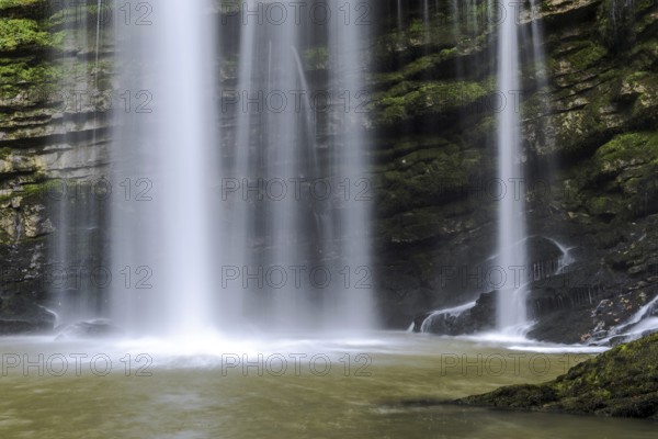 A waterfall with a stream of water flowing out of it. The water is brown and the rocks are green. France Europe