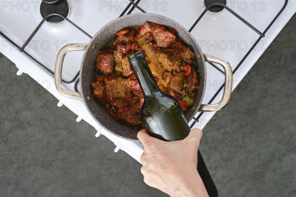 A hand adds a bottle of wine to a pot filled with meat and vegetables simmering on the stove. The cozy kitchen features a gas burner and a modern design, perfect for creating delicious meals