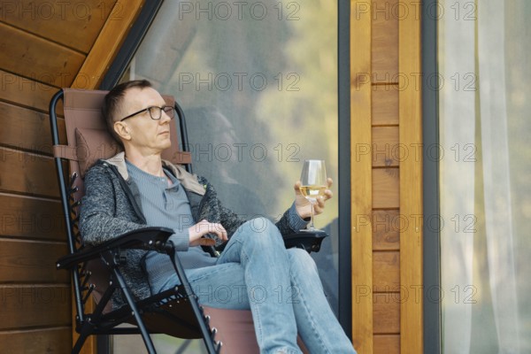 Middle-aged man relax on terrace of a tiny house with a glass of wine