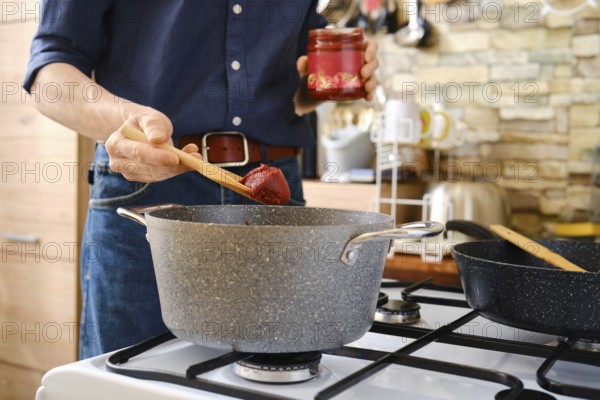 A person prepares a homemade meal adding tomato paste to a pot on the stovetop in a bright, modern kitchen