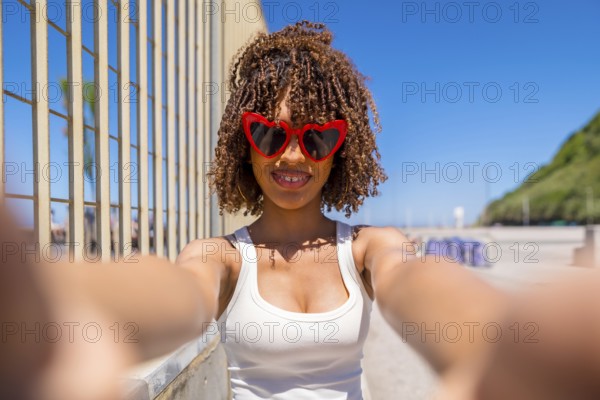 Smiling young woman with curly hair taking a selfie with heart shaped sunglasses on a sunny beach boardwalk