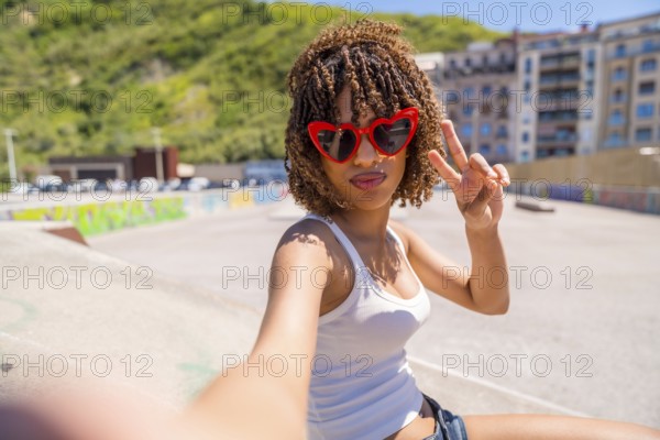 Fashionable young woman with heart shaped sunglasses making peace sign while taking a selfie in skatepark