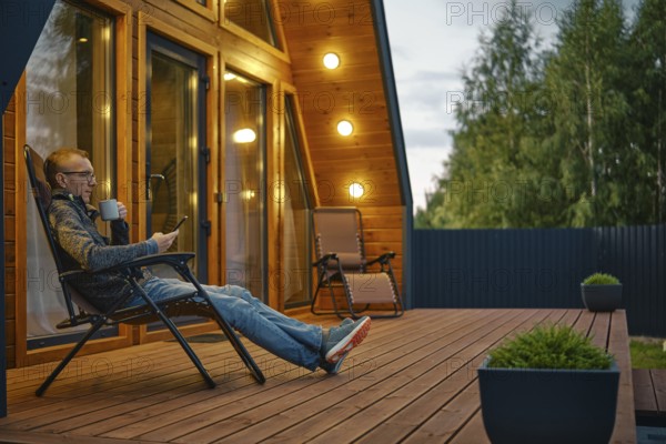 Middle-aged man sits with a cup of tea in the evening on the terrace illuminated by outdoor wall lamps