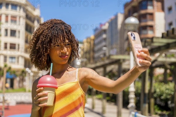 Young woman taking a selfie with her smartphone while holding a milkshake, enjoying a sunny day in the city during her summer vacation