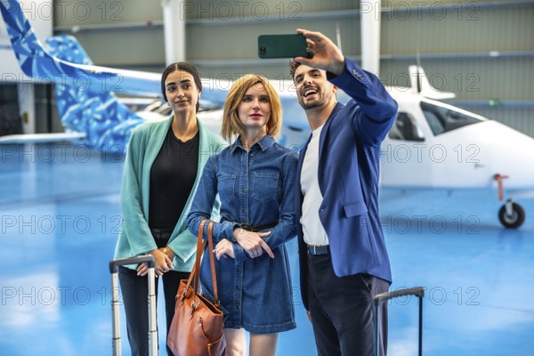 Three businesspeople are taking a selfie with a smartphone in a hangar with a private jet and luggage