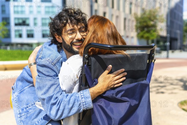 Young man with glasses is embracing a red haired woman sitting in a wheelchair on a sunny day in the city