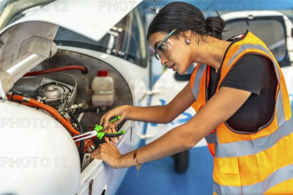 Young woman aircraft mechanic wearing safety glasses and high visibility vest using pliers while repairing airplane engine in hangar