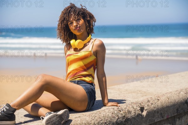 Young woman enjoying music with headphones while relaxing on a beach wall, embracing the summer vibes