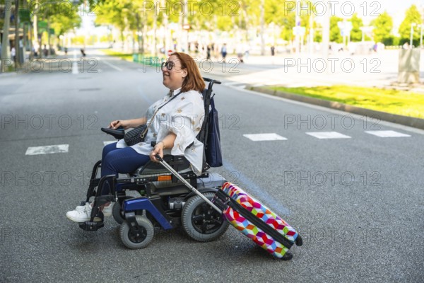 Happy woman with disability enjoys her accessible travel, pulling colorful suitcase while riding modern electric wheelchair on city street