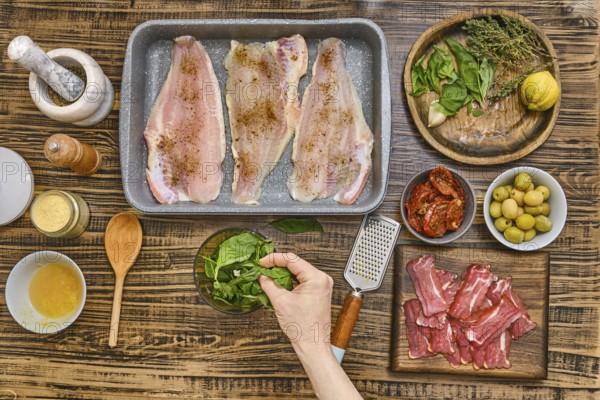 Fresh herbs, vegetables, and fish fillets are arranged on a wooden table. A hand adds spinach to a bowl while spices and garnishes await the next steps in crafting a delicious meal