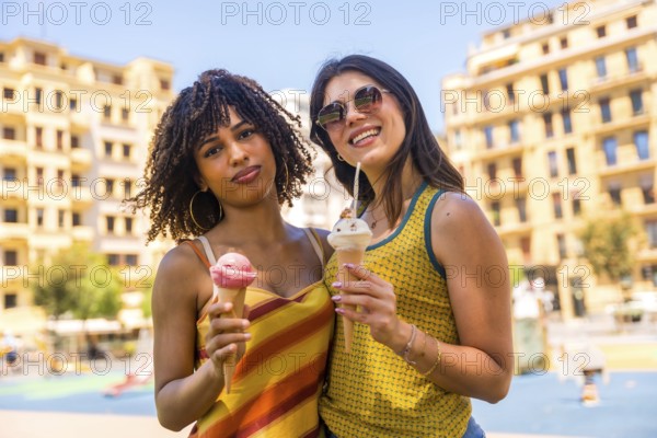 Two female friends enjoying ice cream cones together on a sunny summer day in a city park, sharing a moment of happiness and refreshment