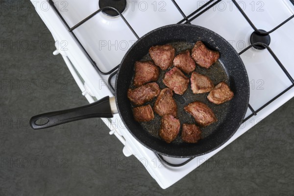Diced beef is sizzling in a black skillet on a white stove. The meat is lightly browned and ready for further cooking. This preparation is taking place in a home kitchen setting around midday