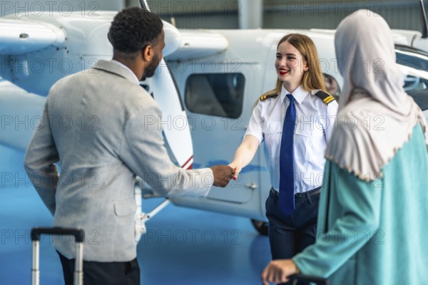 Female pilot shaking hands with businessman next to a private airplane inside an hangar, a businesswoman is waiting next to them