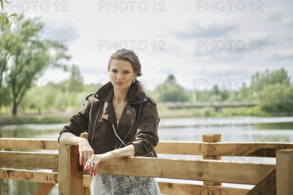 A woman stands confidently by a wooden railing in an elegant outfit, enjoying a peaceful afternoon by the lake with lush greenery and soft clouds in the background