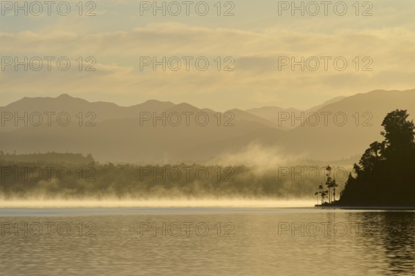 A calm lake at dawn with mist, surrounded by silhouetted mountains, Lake Mahinapua, Ruatapu, South Island, New Zealand, Oceania