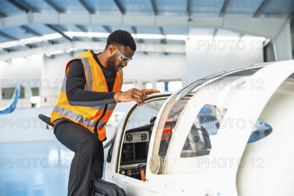 Aircraft maintenance engineer wearing safety glasses and a high visibility vest is inspecting the cockpit of a small plane inside an aircraft hangar