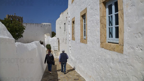 Two people walking along white buildings with windows on a sunny day, Mantomata Stones, Simandiri House, Chora, UNESCO World Heritage Site 1999, Patmos, Dodecanese, Greek Islands, Greece, Europe