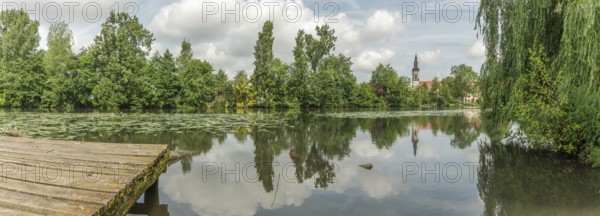 A lake with a tree in the background. The water is calm and still. A building can be seen in the background. Huttenheim, Bas rhin, Alsace, Grand Est, France, Europe