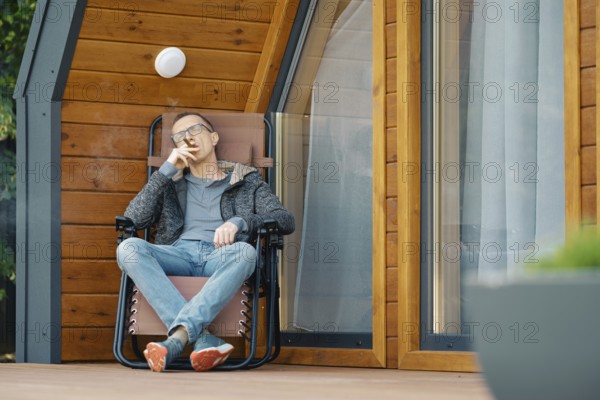 Middle-aged man sitting in lounge chair on the terrace of a modern tiny house with cihgar