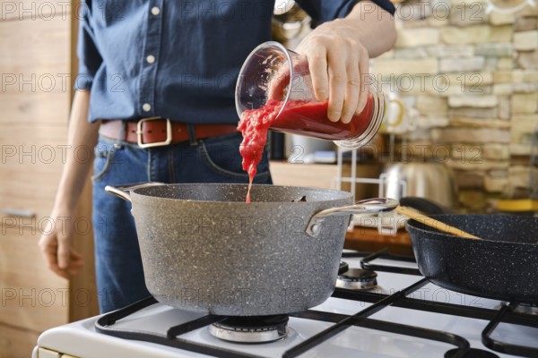 A person pours tomato sauce from a jar into a pot on the stove. The kitchen features modern decor with a warm ambiance