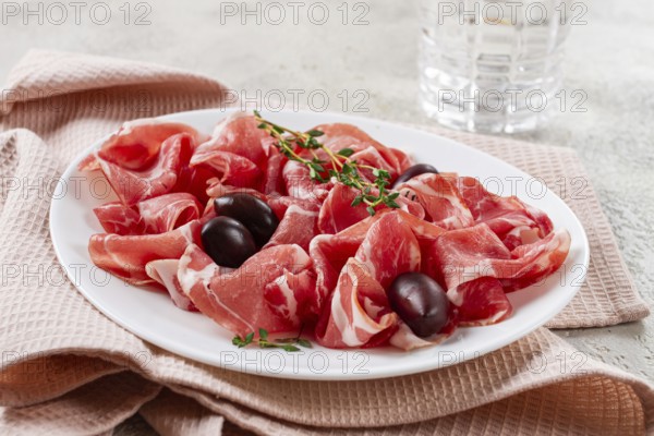 Freshly cut dried meat, served with olives and herbs, on a white plate, light background, no people