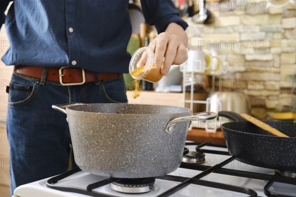 A person pouring spices into a pot on a stove. The countertop is lined with cooking utensils, and a colorful backdrop adds to the homely atmosphere