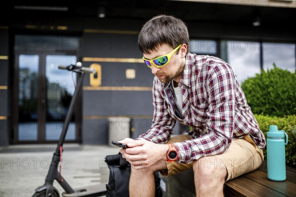 Individual urban environmental friendly transport electric kick scooter parked near man sitting on wooden bench in recreation area. Male use phone, surfing Internet, online shopping at smartphone