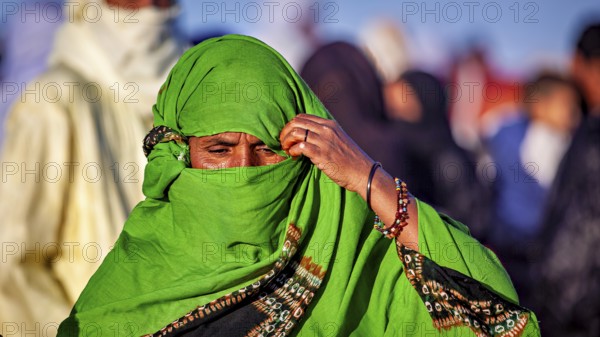 A woman in a green robe and jewellery pensively covers her face, Veiled woman at the Tamanrasett Festival in the Sahara in Algeria