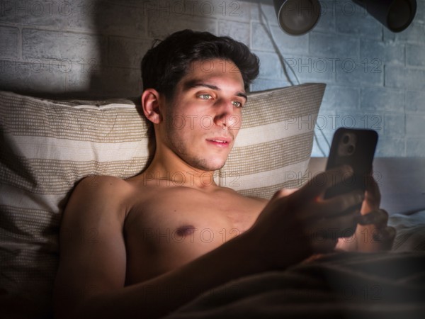 Handsome young man in bed typing on cell phone, sending text message or dialing number