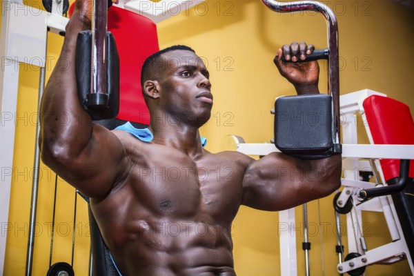 Hunky muscular black bodybuilder working out in gym, exercising pecs on machine