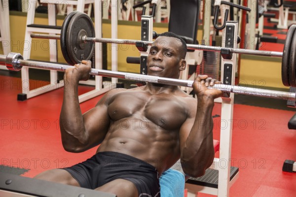Hunky muscular black bodybuilder working out in gym, exercising pecs on bench with barbell