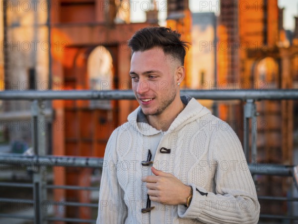 Handsome trendy young man, smiling, standing on a sidewalk in city setting at sunset wearing a fashionable sweater or cardigan