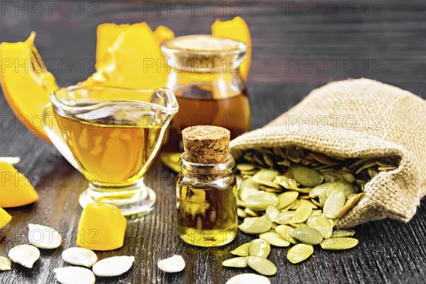 Pumpkin oil in vial, gravy boat and a jar, seeds in a bag and on table, slices of vegetable on black wooden board background