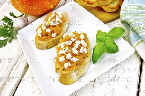 Bruschetta with pumpkin, salted feta cheese in a plate, garlic, basil and parsley, towel and orange vegetable on a wooden board background