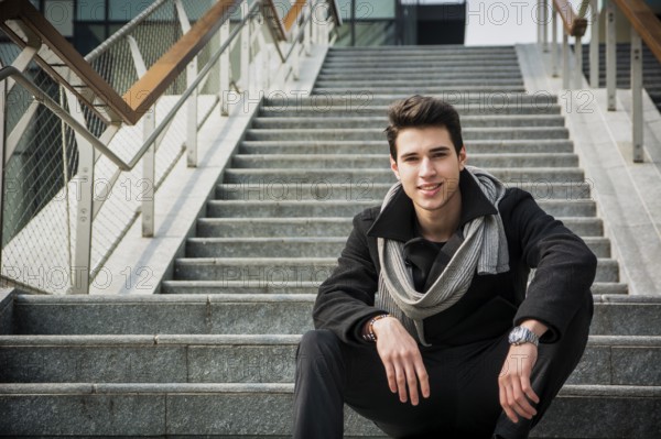 Trendy handsome young man in winter fashion sitting on a long staircase leading to an urban commercial building looking at the camera smiling