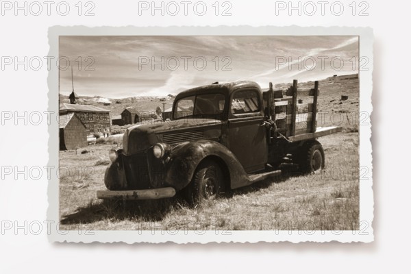 Old lorry rusting in a meadow in the former gold mining town of Bodie. Photo in sepia tone with white jagged edge in the character of old photos and a reclining shadow. Old truck is rusting on the site of the former gold mining town of Bodie. Photo in sepia tone with white jagged edge in the character of old photos