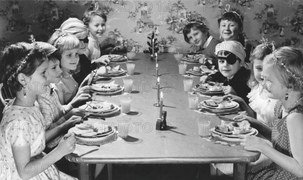 Children in the 1950s. Girls and boys dressed up in hats and costumes are sitting a table together celebrating something, eating cake and having lemonade. Sweden 1957